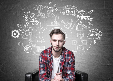 Portrait of a bearded young man sitting in a modern armchair. Blackboard background with a detailed business scheme on it.の写真素材