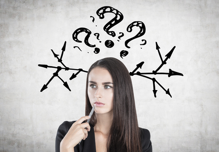 Portrait of a pensive businesswoman with a pen standing near a concrete wall with many question marks above her head and arrows by its sidesの写真素材