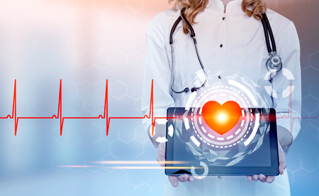 Close up of an unrecognizable woman cardiologist holding a tablet computer with a blank screen. Blurred background with a heart HUD and a cardiogram. Toned image double exposureの写真素材
