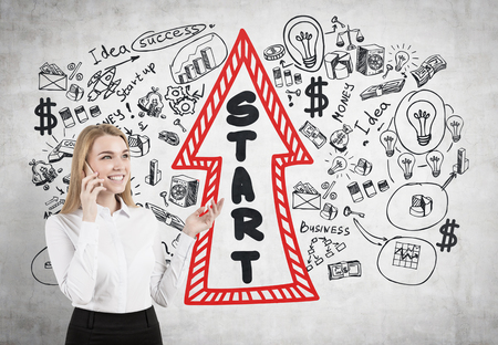 Portrait of a young blonde businesswoman wearing a white shirt and talking on a cell phone and gesticulating. Concrete wall with a start up arrowの写真素材