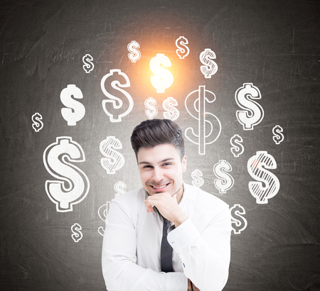 Close up of a cheerful young businessman wearing a white shirt and a tie and sitting near a blackboard with dollar signs drawn on it. Toned imageの写真素材