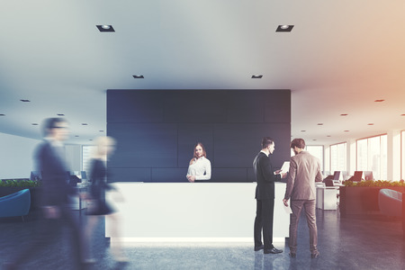 Front view of a white reception desk standing in an open space office environment with a black wall, rows of computer tables and loft windows. People 3d rendering mock up toned imageの写真素材