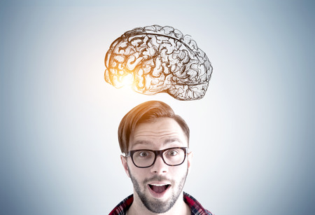 Close up of an astonished young man wearing glasses and standing with an open mouth near a gray wall with a brain sketch drawn on it. Toned imageの写真素材