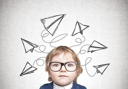 Close up portrait of a cute little boy in a blue business suit wearing glasses and standing near a concrete wall with paper planes drawn above his headの写真素材