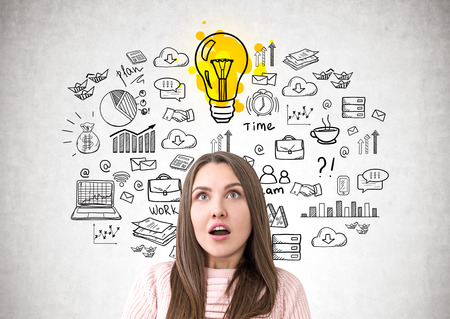 Portrait of an astonished young woman looking upwards. She is wearing a pink sweater and standing near a concrete wall with a business scheme and a light bulbの写真素材
