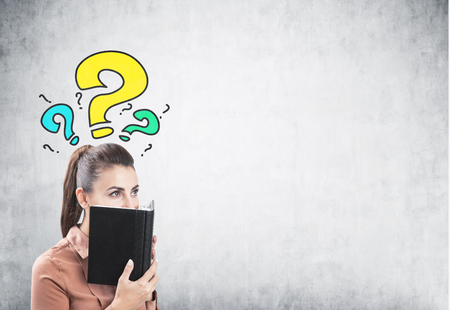 Portrait of a young businesswoman wearing a brown blouse and having long dark hair. She is covering her face with a planner standing near a concrete wall with yellow and blue question marks. Mock upの写真素材