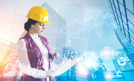 Side view of a young woman wearing a hardhat and looking at documents standing against a cityscape...
