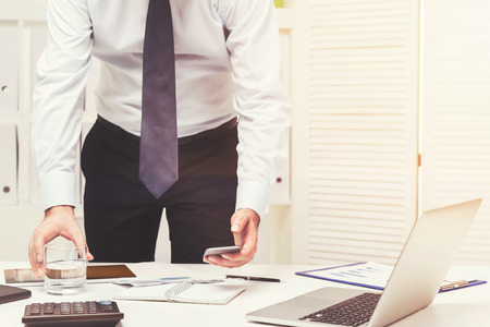 Unrecognizable young businessman is looking at his smartphone standing near an office table. Toned imageの写真素材
