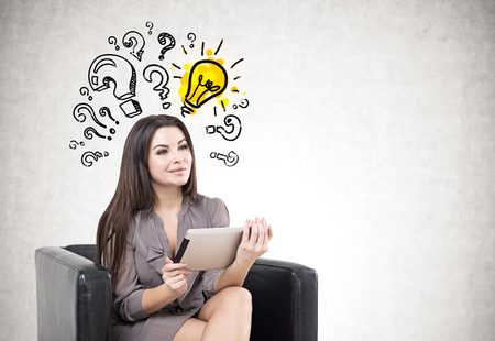 Portrait of a young smiling businesswoman wearing a suit and holding a digital tablet while sitting in an armchair. Gray background with a yellow light bulb and question marks. Mock upの写真素材