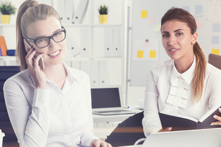 Blonde busiensswoman in glasses is talking on her phone. He colleague with dark hair is holding a planner. Toned imageの写真素材