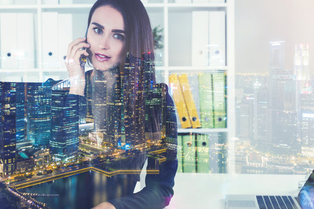 Serious young manager is talking on her phone while sitting at her workplace. There is a night cityscape in the foreground. Toned image double exposureの写真素材