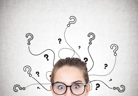 Close up portrait of a young astonished woman wearing round glasses and standing near a concrete wall with many question marks around her headの写真素材