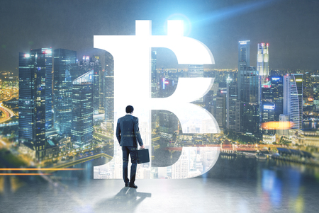 Rear view of a businessman holding a suitcase and looking at a night cityscape standing in a room with a large bitcoin sign opening in the wall. Toned image mock upの写真素材