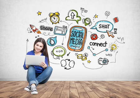 Portrait of a smiling young woman with long dark hair wearing a jeans shirt and sitting on the floor with her laptop near a colorful social media sketch on a concrete wall.の写真素材