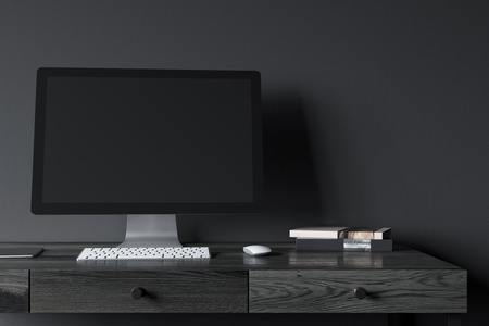 Blank computer screen standing on a black wooden desk in a modern office. A black wall background....