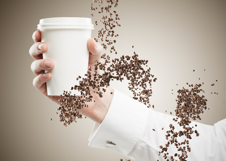 Hand of an unrecognizable businessman wearing a white shirt and holding a paper cup of coffee with coffee beans swirling around him. Mock upの写真素材