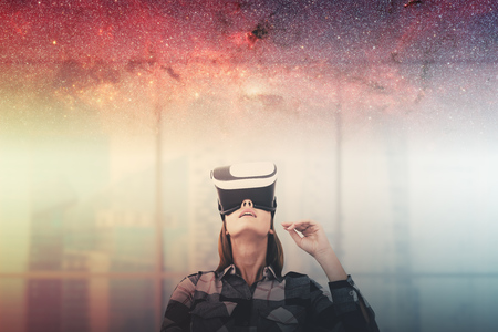 Young woman in a checkered shirt wearing VR glasses and looking upwards. A blurred office background...