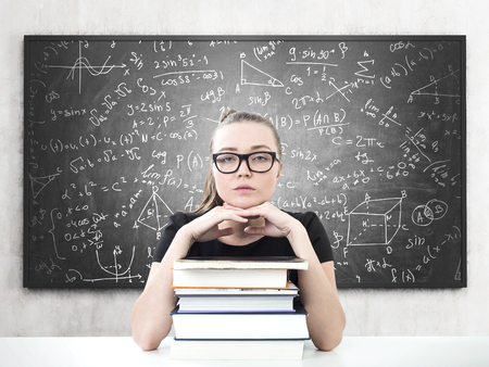 Portrait of a serious young woman in glasses looking forward and sitting with her chin on a stack of books. A blackboard with formula on itの写真素材