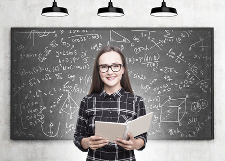 Portrait of a smiling young woman wearing a checkered dress and glasses is holding an open copybook. A blackboard with education iconsの写真素材