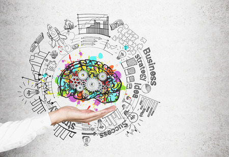 Hand of an unrecognizable businessman holding a colorful brain sketch with cogs on it and a business idea sketch. A concrete wallの写真素材