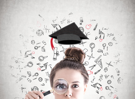 Businesswoman s head with a magnifying glass near her eye. A concrete wall with an education sketch on it.の写真素材