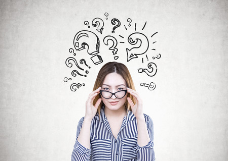 Attractive and serious Asian businesswoman wearing a striped shirt is touching her glasses and looking at the viewer. A concrete wall with question marksの写真素材