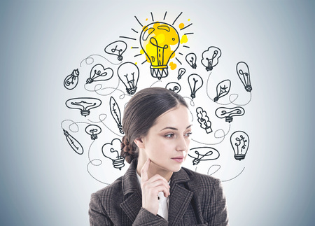 Thoughtful young woman with long dark hair wearing a suit is thinking. She is looking sideways and touching her face with a finger. Yellow light bulbs on a gray wallの写真素材