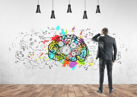 Rear view of a thoughtful well dressed businessman scratching his head. Concept of confusion and a helping hand. A concrete wall background with a cog brain sketch on it.の写真素材