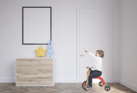 Cute little boy in a white shirt and dark blue jeans is riding a tricycle and showing with his finger. A baby boy room background with a poster above a set of drawersの写真素材