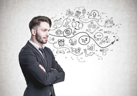 Side view of a pensive young businessman with a beard. He is wearing a suit and a tie and standing with crossed arms. A concrete wall background with a start up sketchの写真素材
