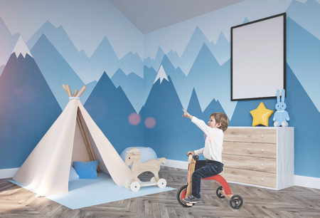 Cute little boy in a white shirt and dark blue jeans is riding a tricycle and showing with his finger. A baby boy room background with a poster mock up and a tentの写真素材