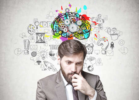 Young businessman with dark hair and a beard wearing a suit is thinking. A gray wall background with a cog brain sketch and business icons on it. Concept of doubt and choice in decision makingの写真素材