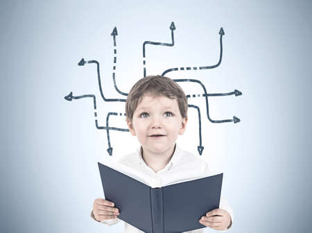 Portrait of a cute little boy in a white shirt holding an open book and looking upwards. A gray wall with an arrows maze sketchの写真素材