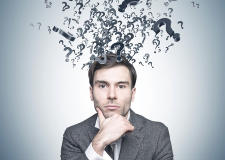 Head and shoulders portrait of a thoughtful young businessman with dark hair looking at the camera. A gray wall background with lots of question marksの写真素材