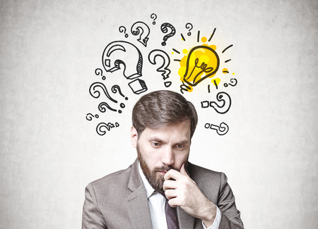 Young businessman with dark hair and a beard wearing a suit is thinking. A concrete wall background with question marks and a yellow light bulb. Concept of doubt and choice in decision makingの写真素材