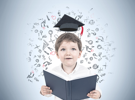Portrait of a cute little boy in a white shirt holding an open book and looking upwards. A gray wall...