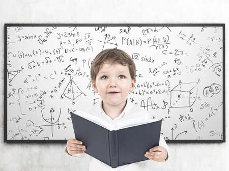 Portrait of a cute little boy in a white shirt holding an open book and looking upwards. A whiteboard background with formulas written on itの写真素材
