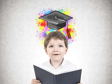 Portrait of a cute little boy in a white shirt holding an open book and looking upwards. A concrete...