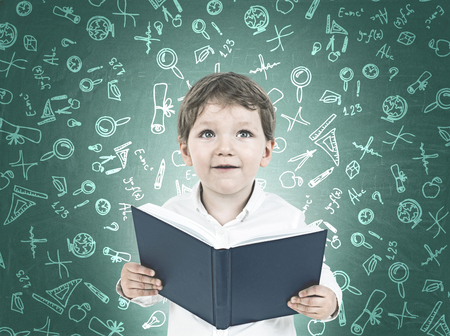Portrait of a cute little boy in a white shirt holding an open book and looking upwards. A green...