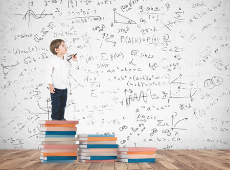Cute little boy in a white shirt and dark jeans holding a marker and looking upwards standing on a book pile. A concrete wall background with formulas written on itの写真素材
