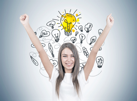 Smiling young woman with brown hair wearing a white t shirt is standing with her hands in the air and looking at the viewer. Yellow light bulbs on a gray wall. An idea conceptの写真素材