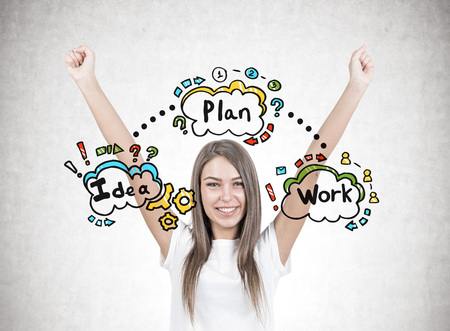Smiling young woman with brown hair wearing a white t shirt is standing with her hands in the air and looking at the viewer. A concrete wall with an idea, plan, work sketchの写真素材