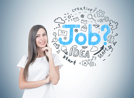 Smiling young woman with fair hair wearing a white T shirt and thinking while looking at the viewer. A gray wall with a round job search sketch on it.の写真素材