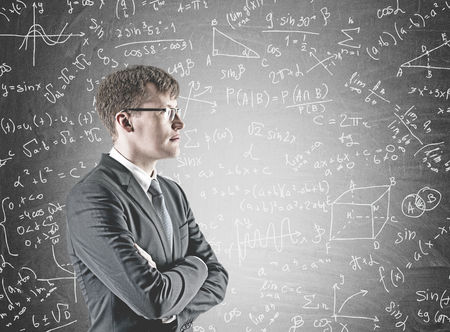 Side view of a confident young businessman wearing a suit and glasses and standing with crossed arms...