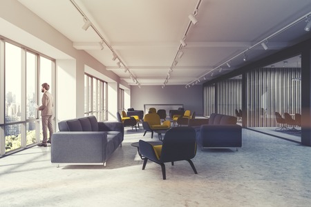 Businessman in a an office waiting room with loft windows, a concrete floor and yellow and blue armchairs standing next to dark blue sofas. 3d rendering toned imageの写真素材