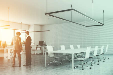 Two businessmen talking in a white conference room of a modern company office with a concrete floor,...