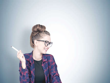 Smiling confident young hipster woman wearing a checkered shirt, a black t shirt and glasses holding a pen and looking sideways. A gray wall background. Mock upの写真素材