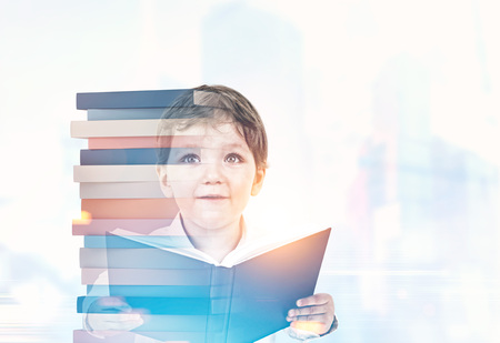 Portrait of a cute little boy in a white shirt holding an open book and looking upwards. A pile of books, a blurred cityscape background. Mock up toned image double exposureの写真素材