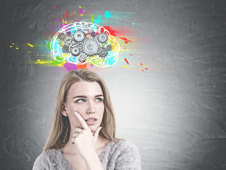 Head and shoulders portrait of a gorgeous young woman with gray eyes and blond hair looking into distance and thinking. A blackboard with a cog brain sketch on it. Mock upの写真素材