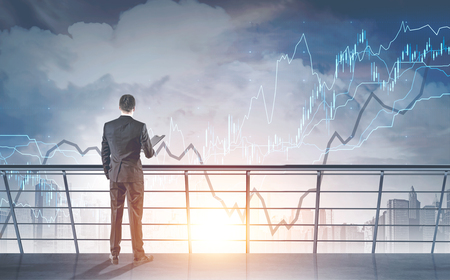 Rear view of a young businessman in a gray suit holding a folder. He is looking at graphs in a morning city sky standing on skyscraper balcony. Toned image double exposure mock upの写真素材